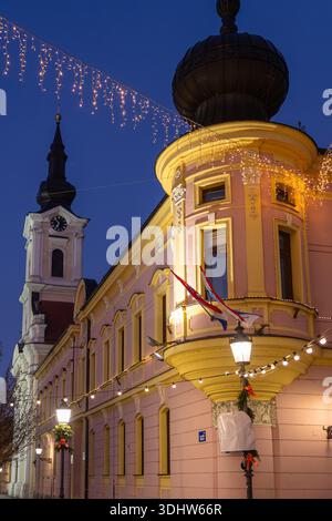 Un'affascinante scena urbana al crepuscolo con un edificio d'angolo rosa, luci a corda, una torre a cupola e bandiere. La luce calda emessa dai lampioni stradali crea un effetto fe Foto Stock