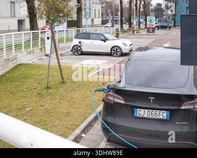 Cremona, Italia 9 gennaio 2026 due veicoli elettrici, una Tesla Model Y e una Mini Cooper se, che si caricano presso una stazione pubblica in un ambiente urbano, rappresentano Foto Stock