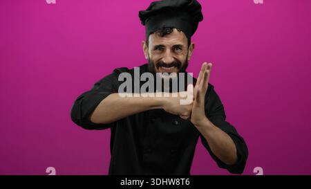 Chef ispanico barbuto che gestiva aggressivamente in uniforme nera su un vivace sfondo rosa che ritrae frustrazione ed emozione. Foto Stock