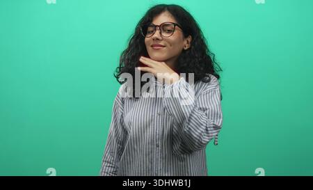 Giovane donna araba con capelli ricci e occhiali che toccano la guancia con mano in studio verde che indossa camicia a righe e colletto abbottonato; calma confida Foto Stock