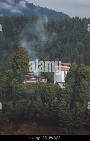Vista di un tradizionale edificio bhutanese con pareti bianche e tetti rossi accoccolati tra lussureggianti alberi verdi, fumo che sale dolcemente nell'aria nebbiosa, Jakar, Foto Stock
