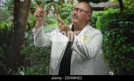 Uomo medico, ispanico anziano nel camice da laboratorio punta il dito verso l'alto con entrambe le mani vicino al passaggio del giardino, costruendo, indossando occhiali e penne tascabili; curiosit Foto Stock