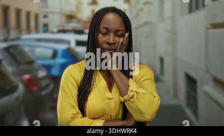 Donna in camicia gialla con capelli intrecciati si stende su strada urbana, esprimendo lo stress con gli occhi chiusi e le braccia incrociate, circondata da auto parcheggiate in un ci Foto Stock