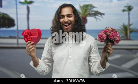 Giovane con i capelli lunghi e la barba che tiene in mano il cuore rosso e il bouquet rosa sulla strada vicino al lungomare con le palme; sorpresa romantica. Foto Stock