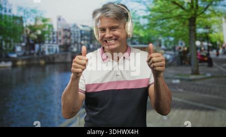 L'uomo con i capelli grigi che indossa le cuffie sulle orecchie sorride e regala due pollici mentre si trova in piedi accanto a un passaggio pedonale sul canale in una strada; felicità e svago. Foto Stock