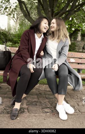 Due donne che sorridono l'un l'altra mentre si siedono insieme sulla panchina del parco, divertenti, divertiti, gioia, unione, amicizia, amico Foto Stock