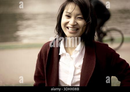 Donne asiatiche felici sorridono gioiosamente come passanti accanto a un ciclista che passa sul marciapiede vicino a un litorale, trasudando felicità in una giornata piacevole Foto Stock