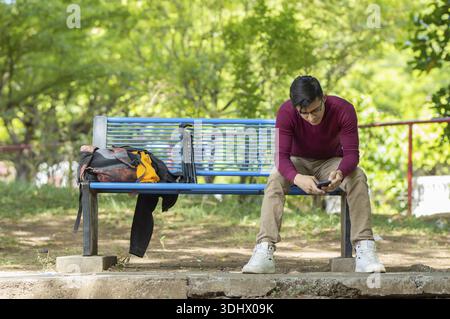 Giovane uomo con i suoi effetti personali seduto su una panchina del parco. Uomo che guarda il telefono con lo zaino accanto a lui, seduto nel parco Foto Stock