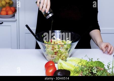 Persona che prepara sana insalata verde, versando condimento con olio d'oliva su lattuga, avocado e pomodoro in una ciotola di vetro sul bancone della cucina Foto Stock