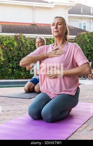 La coppia senior si inginocchia esercitandosi a respirare indossando abiti da ginnastica su tappeti viola e scuri a bordo piscina Foto Stock