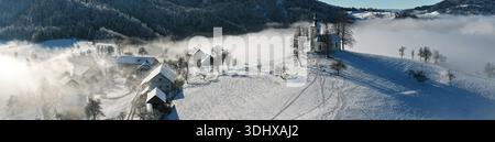 Panorama invernale ad alta risoluzione della Chiesa di Jamnik nella nebbia mattutina, vista aerea del Villaggio Sloveno nelle Alpi Giulie, paesaggio mistico di St prim Foto Stock
