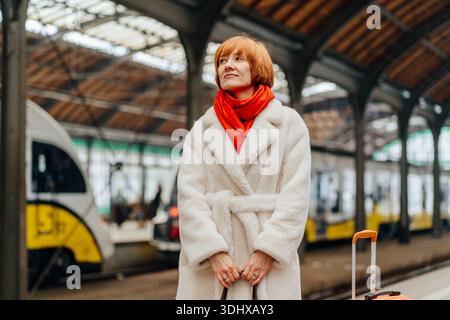 Donna matura e sicura di sé con i capelli rossi in piedi sulla piattaforma della stazione ferroviaria con i bagagli Foto Stock