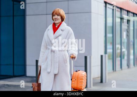Donna matura e sicura che cammina con la valigia in una città moderna Foto Stock