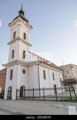 Una tranquilla chiesa bianca con un alto campanile e una croce sorge accanto ad una recinzione nera in ferro battuto in una tranquilla strada urbana. La luce del giorno illumina il clas Foto Stock