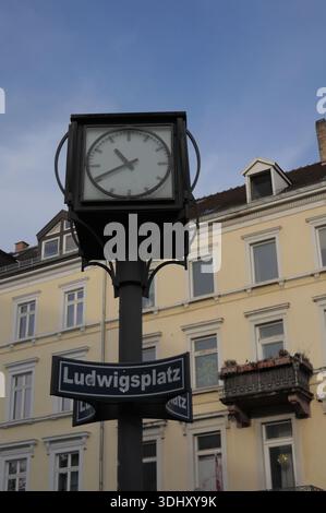 Orologio pubblico montato su un palo nero con un cartello per Ludwigsplatz. Edifici residenziali storici con balconi e facciate dai colori chiari Foto Stock