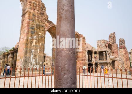 Delhi, India - 30 marzo 2024: L'immagine mostra una vista ravvicinata della colonna di ferro dietro una recinzione metallica, con intricati archi in pietra scolpiti e antiche rovine visibili sullo sfondo del complesso Qutub Minar. Foto Stock