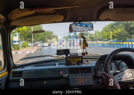 Calcutta, India - 24 marzo 2024: L'immagine mostra l'interno di un taxi Ambassador con la mano del conducente sul volante, i dettagli del cruscotto, il metro di tariffa, l'ornamento appeso e la vista attraverso il parabrezza di un'ampia strada urbana alberata con barriere blu e bianche ai bordi della strada a Calcutta. Foto Stock