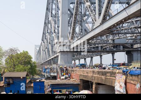 Calcutta, India - 24 marzo 2024: Veicoli e pedoni attraversano la struttura in acciaio del ponte Howrah con edifici in metallo ondulato blu e attività di mercato visibili sotto. Foto Stock