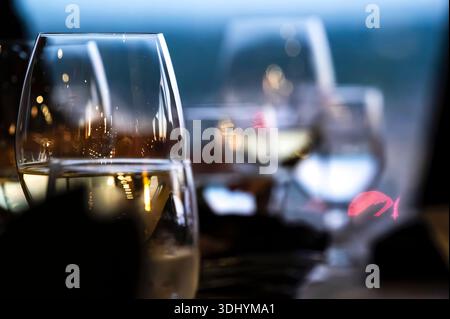 Vino bianco e bicchieri d'acqua si siedono su un tavolo del ristorante, riflettendo la luce dell'ambiente e i delicati colori della città. Il bokeh rosa e viola sullo sfondo suggerisce Foto Stock