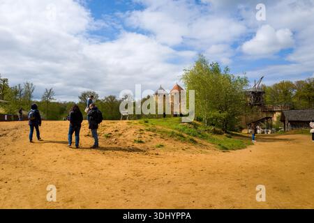 Chateau de Guedelon, Francia - 20 aprile 2024: Si vedono diverse persone camminare e stare in piedi su un sentiero sterrato con le torri di pietra, la gru in legno e il verde circostante dello Chateau de Guedelon visibile sullo sfondo sotto un cielo parzialmente nuvoloso. Foto Stock