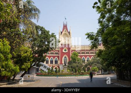 Calcutta, India - 24 marzo 2024: La facciata della Calcutta High Court è visibile dietro alberi frondosi, con un solo pedonale che cammina sulla strada di fronte allo storico edificio rosso e crema sotto un cielo limpido. Foto Stock