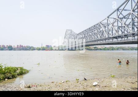Calcutta, India - 24 marzo 2024: Due persone si vedono fare il bagno nel fiume Hooghly vicino alla riva con la struttura in acciaio del ponte Howrah che attraversa il fiume a Calcutta. Foto Stock