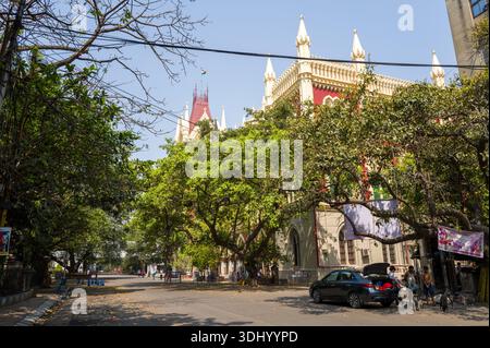 Calcutta, India - 24 marzo 2024: L'edificio della Corte Suprema di Calcutta con la sua architettura gotica rossa e crema è parzialmente visibile dietro alberi frondosi lungo una strada tranquilla, con auto parcheggiate, pedoni e striscioni visti in primo piano sotto un cielo limpido. Foto Stock