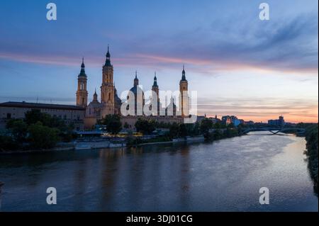 Saragozza, Spagna - 11 maggio 2024: L'immagine mostra la basilica di Nuestra Senora del Pilar con le sue torri e cupole illuminate al tramonto, vista dall'altro lato del fiume Ebro con alberi e un ponte sullo sfondo. Foto Stock