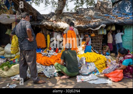 Calcutta, India - 24 marzo 2024: Si vedono persone che vendono e acquistano ghirlande di fiori di arancia e di calendula gialle in un mercato all'aperto con bancarelle, cestini e coperture per teloni improvvisati a Calcutta. Foto Stock