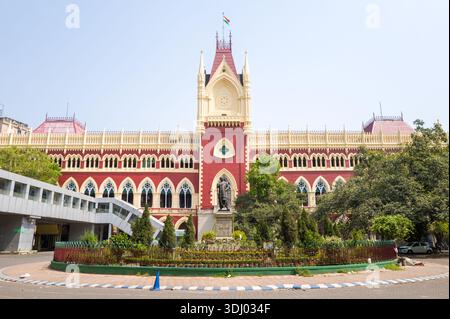 Calcutta, India - 24 marzo 2024: La facciata frontale della Calcutta High Court è visibile con la sua architettura gotica rossa e crema, il giardino centrale con statua e la bandiera nazionale indiana che sventola in cima all'edificio sotto un cielo limpido. Foto Stock