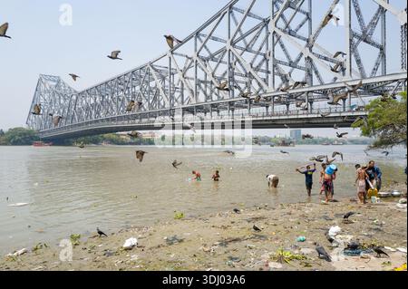 Calcutta, India - 24 marzo 2024: Si vedono diverse persone fare il bagno e lavarsi sulle rive fangose del fiume Hooghly con la struttura in acciaio del ponte Howrah e numerosi uccelli volanti visibili sullo sfondo. Foto Stock
