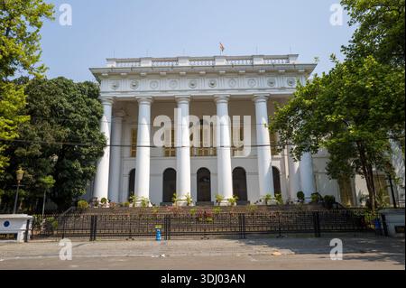 Calcutta, India - 24 marzo 2024: La facciata del municipio di Calcutta, uno storico edificio neoclassico bianco con alte colonne e una bandiera indiana sul tetto, si vede dietro una recinzione di ferro nera e circondato da alberi. Foto Stock