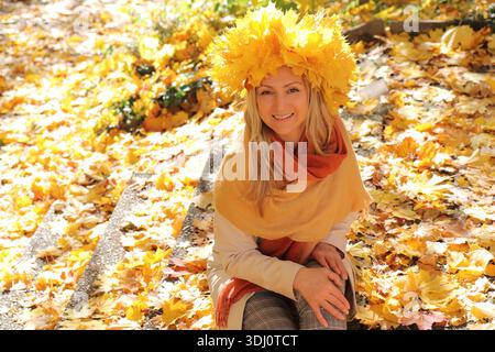 Donna sorridente in una corona di foglie d'acero gialle seduta su scale di pietra ricoperte di fogliame autunnale. Ritratto caldo e soleggiato in un parco stagionale. Foto Stock