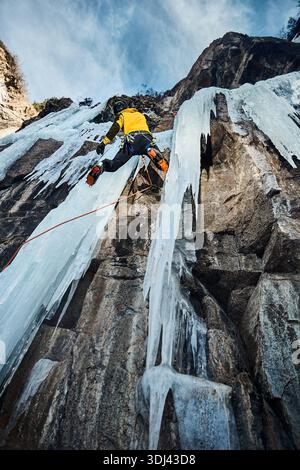 L'uomo con la giacca gialla si arrampica sulla parete di ghiaccio presso la grande cascata ghiacciata con ascia e ramponi contro lo sci blu nelle montagne invernali. Sport alpinista Foto Stock