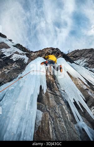 L'uomo con la giacca gialla si arrampica sulla parete di ghiaccio presso la grande cascata ghiacciata con ascia e ramponi contro lo sci blu nelle montagne invernali. Sport alpinista Foto Stock
