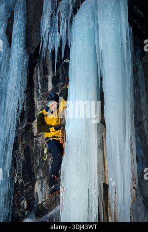 L'uomo con la giacca gialla si arrampica sulla parete di ghiaccio presso la grande cascata ghiacciata con ascia e ramponi contro lo sci blu nelle montagne invernali. Sport alpinista Foto Stock