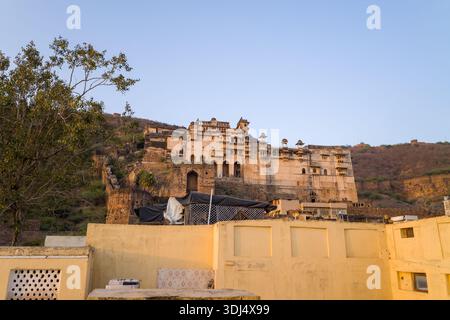 Bundi, India - 19 marzo 2022: Il complesso del Garh Palace è visto su una collina con pareti gialle intemprate, ingressi ad arco e più padiglioni a cupola, con tetti in primo piano e un albero sotto un cielo limpido. Foto Stock