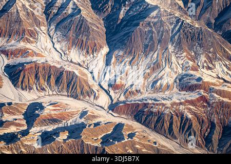 Spettacolare vista aerea delle cime innevate e delle profonde creste tettoniche al tramonto, che passano negli aridi altopiani desertici dell'Asia centrale. Foto Stock