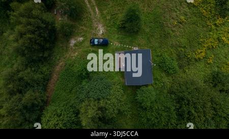 La vista aerea mostra una cabina moderna con tetto scuro, ponte in legno e antenna satellitare in un bosco rurale. Parcheggi bui su una pista sterrata. Fine estate da Foto Stock