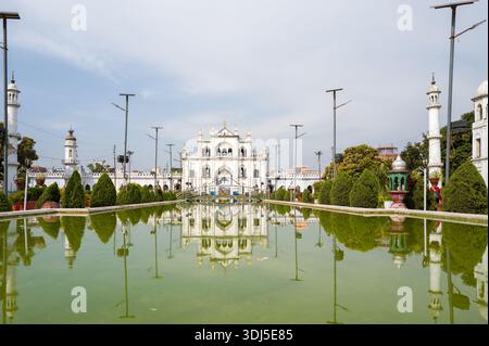 Lucknow, India - 16 marzo 2024: L'immagine mostra il mausoleo Chota Imambara con cupole bianche e minareti, riflessi in una piscina verde con giardino con cespugli rifiniti e lampioni sotto la luce del giorno. Foto Stock