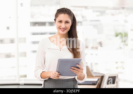 Il tablet viene tenuto a livello del torace in ufficio vicino a grandi finestre, laptop e documenti visibili. Professionale, aziendale, business, spazio di lavoro, moderno, giorno Foto Stock