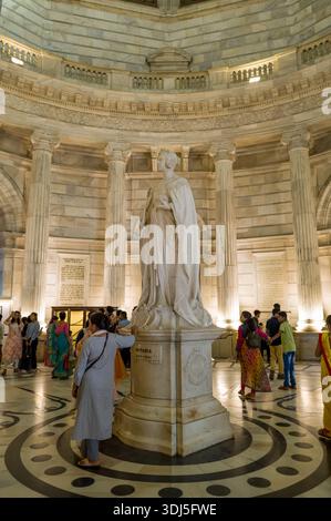 Calcutta, India - 23 marzo 2024: L'immagine mostra un gruppo di persone in piedi e che osservano la grande statua in marmo della regina Vittoria, esposta in modo prominente nella rotonda centrale del Victoria Memorial con colonne neoclassiche e dettagliate pietre visibili. Foto Stock