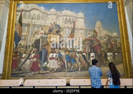 Calcutta, India - 23 marzo 2024: Due persone si trovano di fronte a un grande dipinto incorniciato che raffigura una processione di elefanti reali con cavalieri e assistenti, esposto su una parete interna del Victoria Memorial. Foto Stock