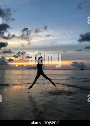 Silhouette di una donna che salta in acque poco profonde sulla spiaggia, formando una forma a cuore con le mani contro il cielo del tramonto. Amore, libertà, felicità. Foto Stock