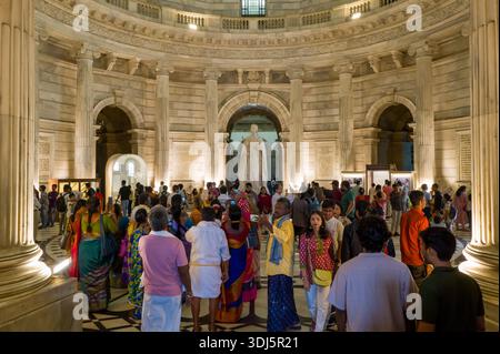 Calcutta, India - 23 marzo 2024: Un grande gruppo di persone si riunisce e cammina intorno alla statua di marmo della regina Vittoria nella rotonda centrale del Victoria Memorial, circondata da colonne neoclassiche e dettagliate pietre. Foto Stock