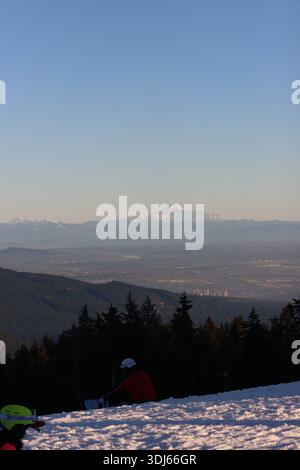 Vista della lontana e accidentata vetta innevata del Monte Baker che si innalza sopra le colline boscose. In primo piano, uno snowboarder siede sulla pista. Foto Stock