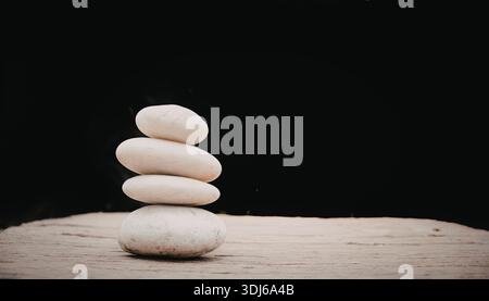 Una pila di quattro pietre bianche bilanciate su una superficie di legno su uno sfondo scuro. L'immagine evoca un senso di calma ed equilibrio. Foto Stock