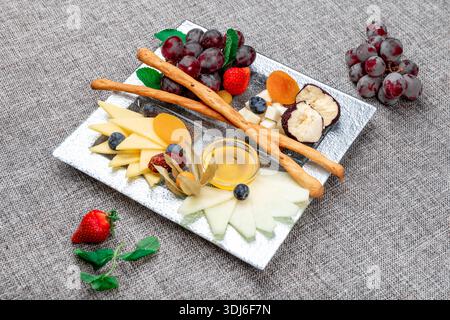 Un vassoio di formaggi gourmet splendidamente organizzato con vari tipi di formaggio a fette, uva rossa, fragole fresche, mirtilli, fisti, e dri Foto Stock
