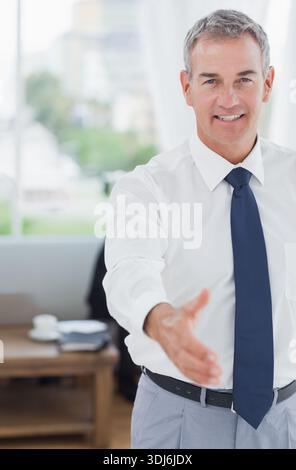 Uomo di mezza età che offre una stretta di mano, indossa camicia bianca e cravatta in ufficio con carte da scrivania. Professionale, executive, illuminato dal sole, finestra, spazio di lavoro, moderno Foto Stock