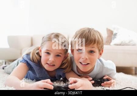 Fratelli e sorelle sdraiati su uno spesso tappeto di colore chiaro a casa, con controller cablati. Fratelli, gaming, accogliente, retroilluminato, Highkey, candido, giorno Foto Stock